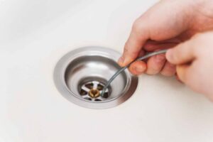 Hand inserting a metal drain snake into a sink drain opening to clear clogs and debris.
