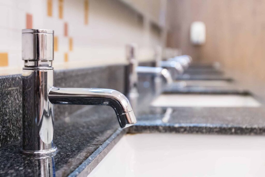Row of polished chrome bathroom sink faucets mounted on dark granite countertops in a commercial restroom setting.