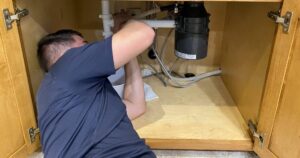 Professional plumber working under sink installing plumbing pipes and black drain canister in wooden cabinet.
