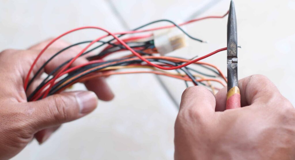 Electrician's hands holding a bundle of colored electrical wires while using a knife tool to strip insulation from a red wire.