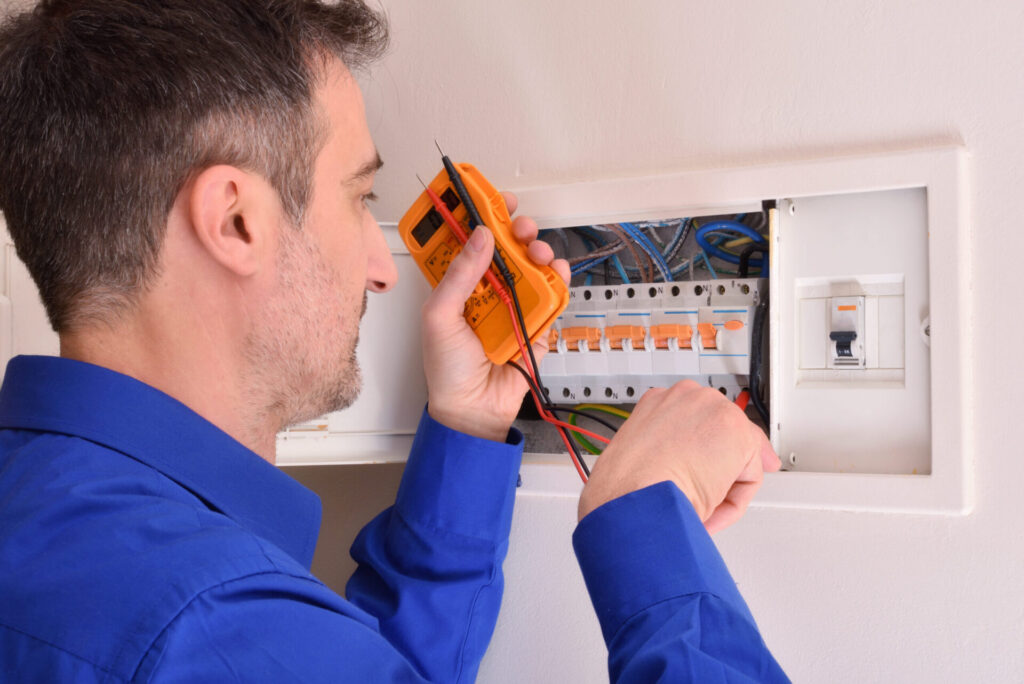 Electrician in blue uniform uses orange multimeter to test electrical panel with orange circuit breakers and colored wiring.