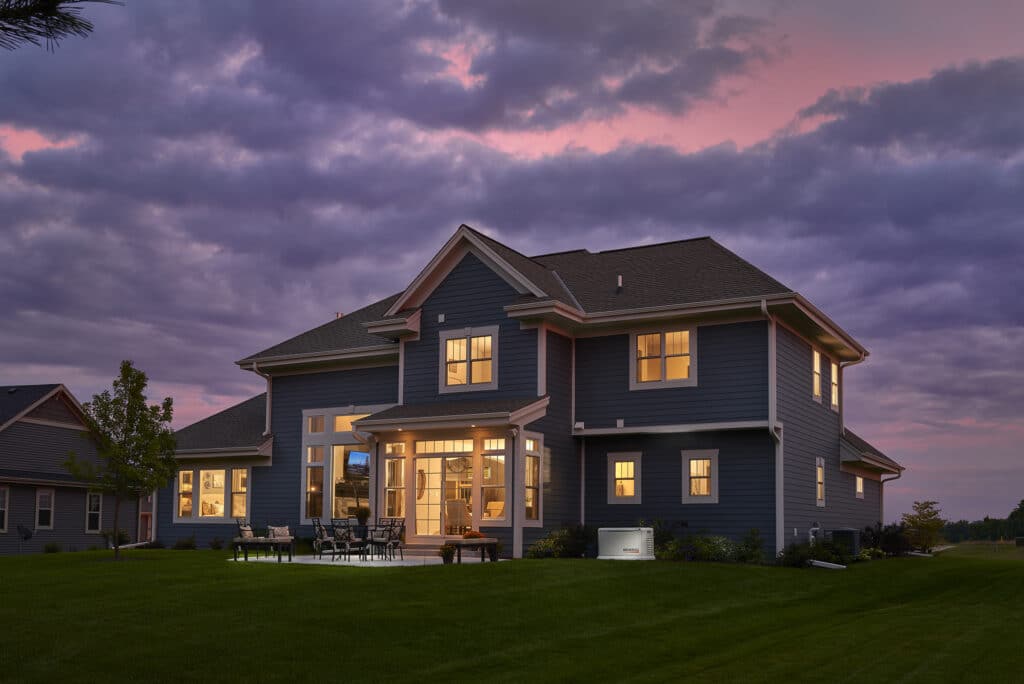 Two-story dark blue residential home illuminated at dusk with a backup generator unit visible in the yard.