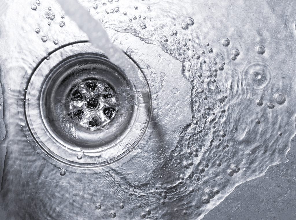 Water flowing down a drain opening with bubbles and ripples visible on the wet metal surface.