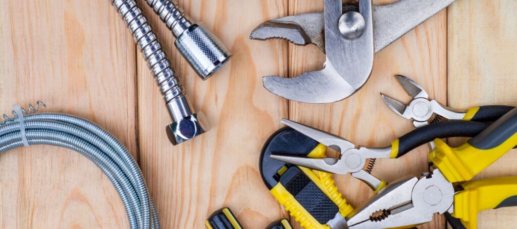 Assorted plumbing tools arranged on wooden surface including wrenches, metal pipe fittings, flexible hose, and needle-nose pliers with yellow handles.
