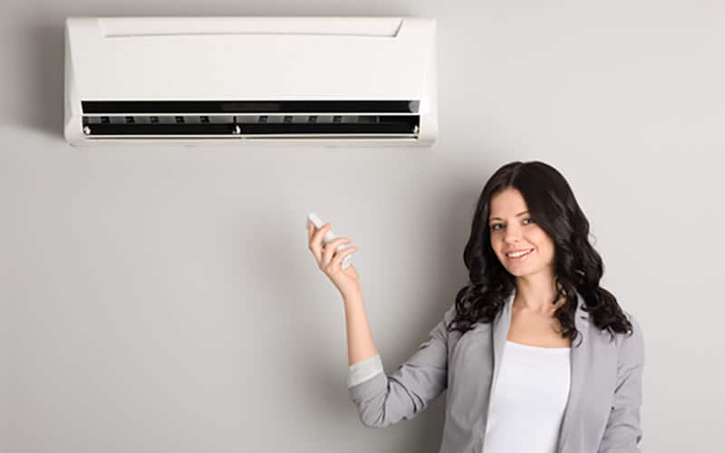 Woman in gray blazer holding remote control toward white ductless mini split air conditioning unit mounted on wall.
