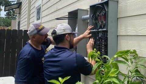 Two technicians in navy uniforms and white caps working on an outdoor electrical panel mounted on a house exterior wall.