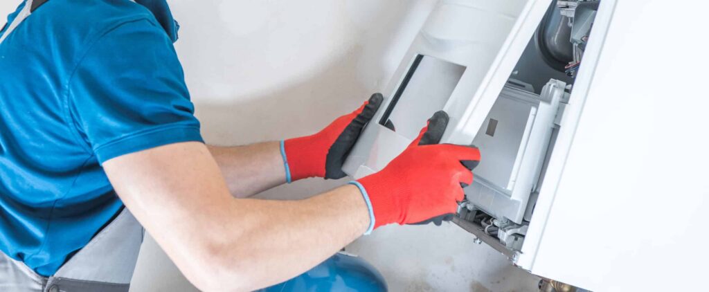 Technician in blue shirt and red work gloves installing an indoor heating unit component during HVAC maintenance.