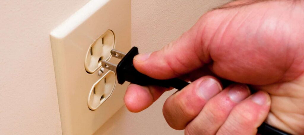 Hand holding a black voltage tester probe inserted into a beige electrical outlet to check if power is present.