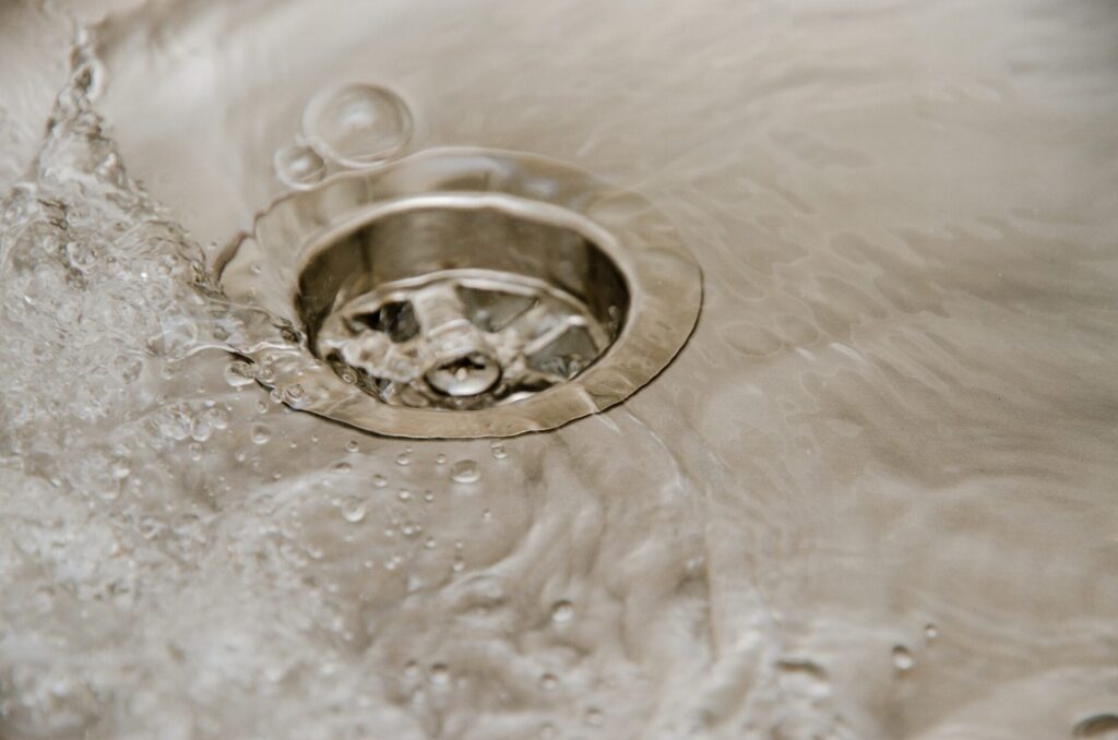 Close-up of a chrome kitchen sink drain with water splashing and droplets on the surrounding stainless steel surface.