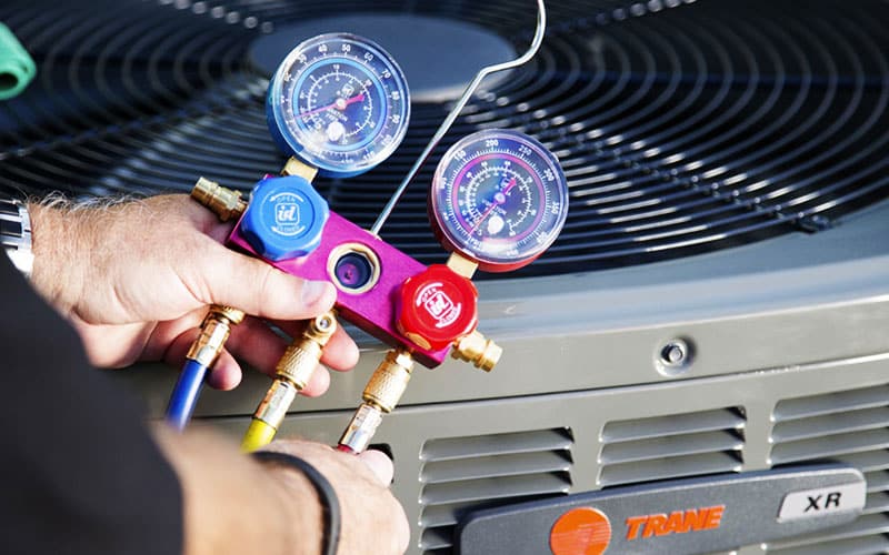 Technician holding red and blue refrigerant manifold gauge set with dual pressure gauges against TRANE air conditioning unit.