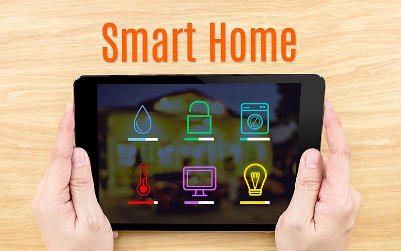Hands holding tablet displaying smart home icons including water, security lock, washing machine, temperature, monitor, and lighting controls.