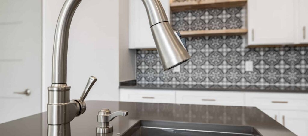 Stainless steel pull-down kitchen faucet with matching soap dispenser above dark countertop sink in contemporary kitchen.