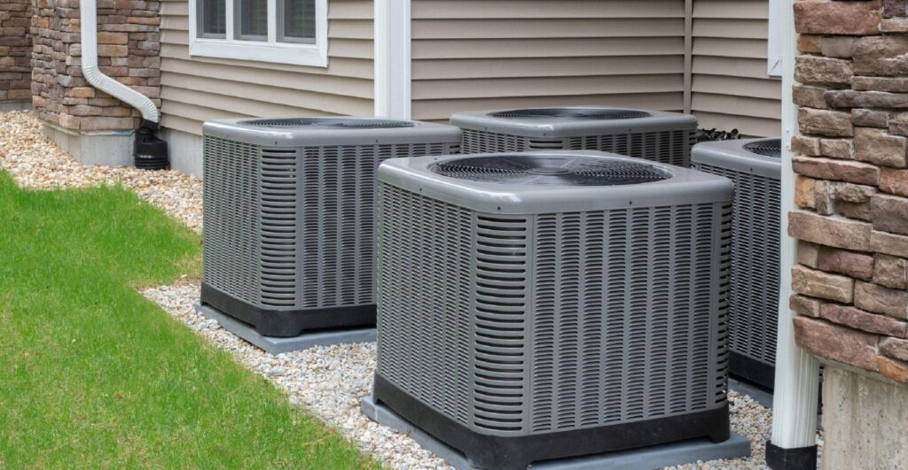 Several gray air conditioning and heating units installed on gravel beside a brick and siding residential home with white trim.