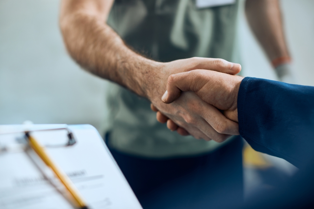 Two men shaking hands over a service agreement clipboard with pen, representing a professional business contract or consultation.