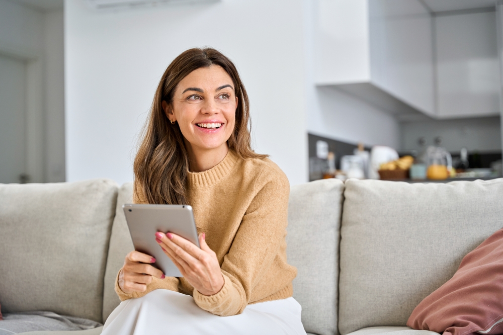 Woman sitting on beige couch in modern home interior, smiling while holding tablet device with kitchen visible in background.