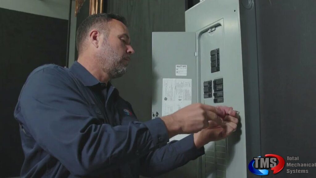 TMS technician in blue work shirt examining an indoor electrical panel with circuit breakers and safety labels.