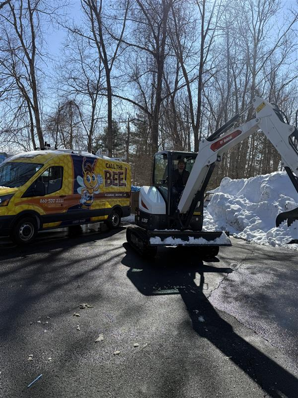 Yellow Call The Bee service van parked next to white excavator removing snow from pavement on sunny day.